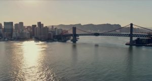 City skyline and a large suspension bridge crossing a reflective harbor at sunset.